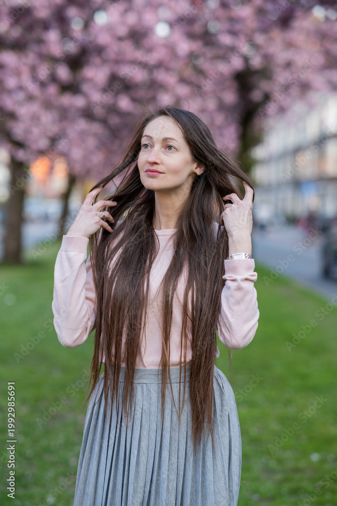 Naklejka premium Woman with long brown hair wearing sunglasses poses among blooming sakura cherry blossom trees in a vibrant spring garden, soft focus background enhancing floral colors