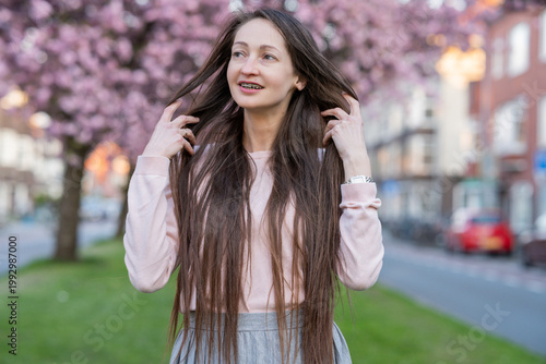 Woman with long brown hair wearing sunglasses poses among blooming sakura cherry blossom trees in a vibrant spring garden, soft focus background enhancing floral colors