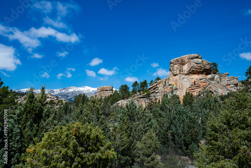 Wide landscape view of La Pedriza featuring a granite rock formation with a barely visible climber in the distance, emphasizing scale and vastness. Surrounded by forest and with a snow-capped mountain