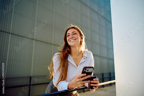 Foto Young woman smiling and holding a mobile phone with a coffee cup, enjoying a bri
