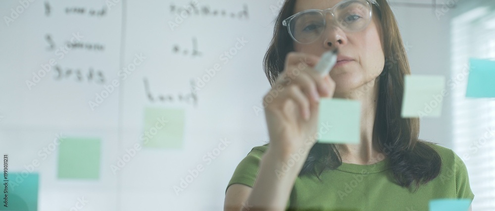 Obraz premium Businesswoman writing notes on glass board, surrounded by sticky papers during strategic planning meeting in sunlit corporate workspace