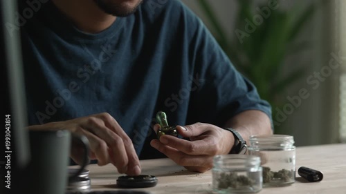 Man filling glass pipe with cannabis, with cannabis equipment and accessories on the table.