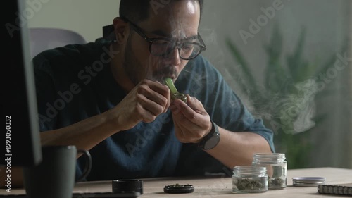 Man smoking a cannabis from smoking pipe, with cannabis equipment and accessories on the table.
