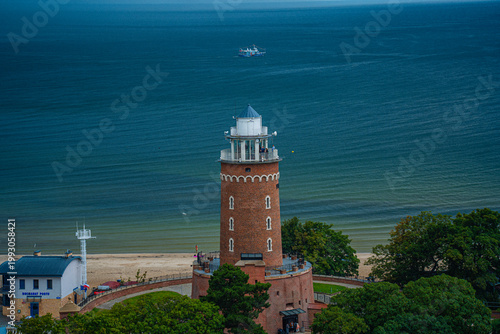 Aerial view of Kołobrzeg historic red brick lighthouse on Baltic Sea coast in Poland, with sandy beach, green trees and fishing boat on calm water.