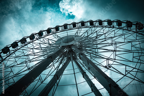 Dramatic low-angle view looking up at large Ferris wheel with gondola cabins against a moody teal dark cloudy sky, geometric steel structure detail.