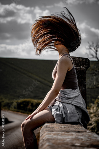 Young woman sitting on stone wall with long hair dramatically blowing in strong wind against moody cloudy sky and green rural hillside landscape.