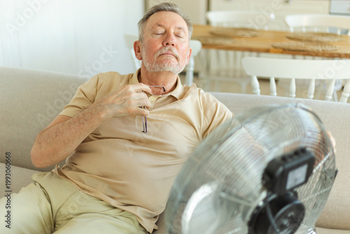 Senior man refreshing himself near fan during summer heat wave hot day. Elderly man suffering from summer heat trying to survive with electric fan. Man enjoying refreshing air blowing from ventilator