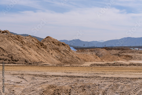 Wide construction site with excavated soil, leveled ground, and distant hills under clear sky, showing early stage land preparation for infrastructure development project.