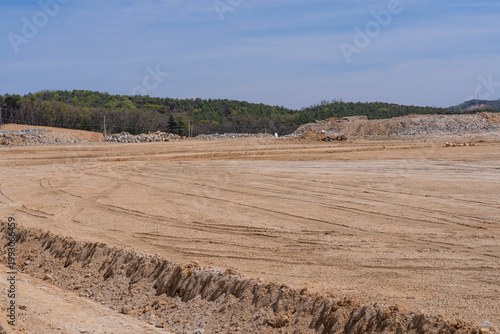 Wide construction site with excavated soil, leveled ground, and distant hills under clear sky, showing early stage land preparation for infrastructure development project.