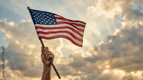 A persons hand firmly holds the American flag against a beautiful sky with golden clouds and sun rays at sunset