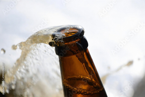 A bottle of beer is being opened, creating a spray of bubbles. It appears to be outdoors on a cloudy day.