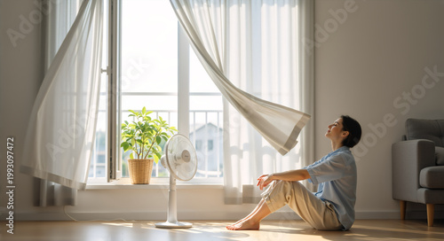 Woman sitting on the floor cooling down with an electric fan. Female relaxing at home by an open window during a summer heatwave