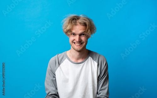 Young man with messy blond hair and a mischievous grin, posing for a cropped studio headshot against a blue backdrop, playful, confident and candid expression