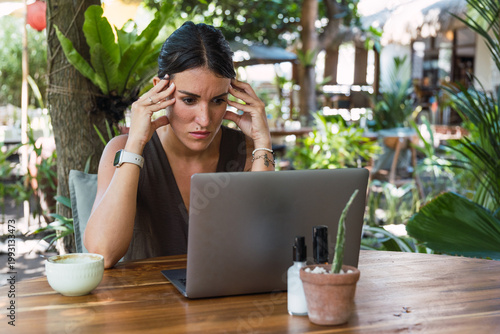Woman focusing on laptop with hands on temples, feeling frustrated at outdoor cafe