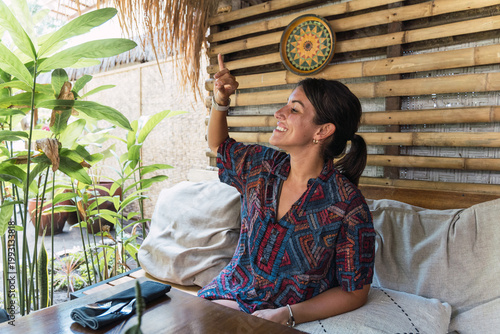 Woman smiling, sitting in a tropical cafe, pointing up to get attention