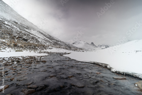 A minimalist winter landscape of Zero Point in North Sikkim, featuring a dark, winding river cutting through a vast, pristine expanse of pure white snow under high-altitude Himalayan conditions.