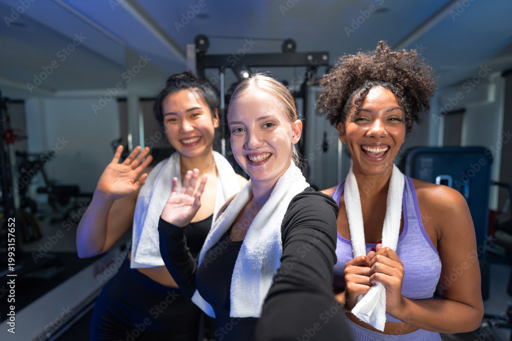Fototapeta premium Cheerful multiethnic friends wave and pose for a post workout selfie in a modern gym with blue mood lighting.