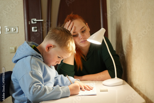 Young boy writing homework while mother supervises at home desk. Education, concentration and parenting concept.