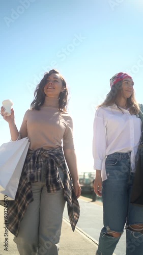 Two stylish young female friends walking along a sunny promenade carrying shopping bags and coffee
