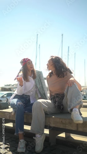 Two smiling girlfriends sitting on a bench in a harbor, chatting and enjoying a beautiful sunny day
