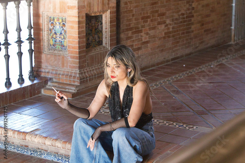 Obraz na płótnie A young, attractive woman wearing jeans, gloves and a black T-shirt, smoking a cigarette whilst sitting on the steps of Spain Square in Seville