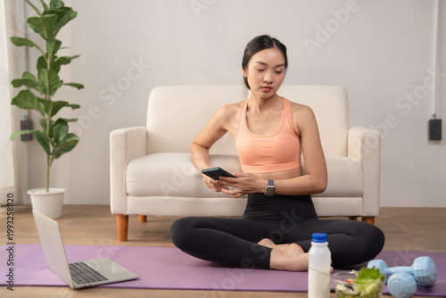 Free Download Young Asian woman sitting on yoga mat using smartphone before home workout, preparing for online fitness training with laptop, weights, milk, and salad - GFXLoad