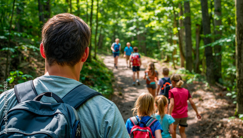 Camp counselor leading a group of children with backpacks on a hiking trail through a lush green forest during summer camp