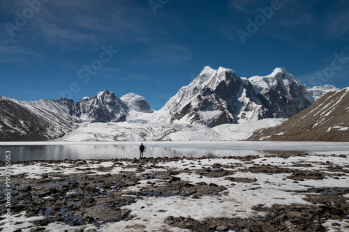 A solitary man stands in the vast, desolate landscape of Gurudongmar, North Sikkim, gazing toward snow-capped Himalayan peaks under a dramatic sky.