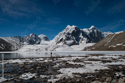A solitary man stands in the vast, desolate landscape of Gurudongmar, North Sikkim, gazing toward snow-capped Himalayan peaks under a dramatic sky.
