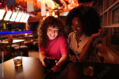 Diverse friends laughing, connecting, viewing content on smartphone at bar