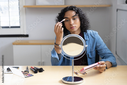 Non-binary person wearing denim shirt applying eyeshadow with brush at light wood table with mirror