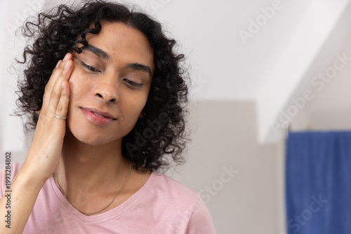 Non-binary person touching left cheek and looking down in bathroom with pink shirt, chain, ring