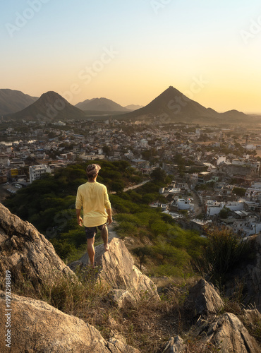 Man standing on a rocky viewpoint overlooking Pushkar, Rajasthan, India, at sunrise with warm light over the town and surrounding hills