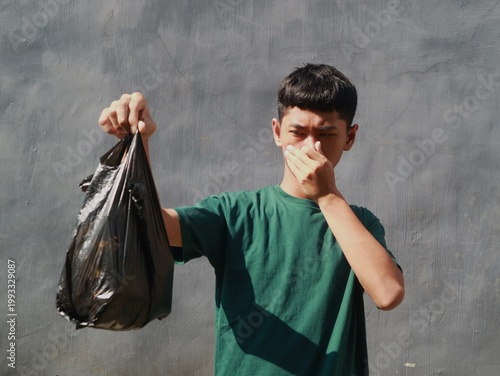 Photos A young man with a disgusted face holds a black plastic bag in front of a gray w