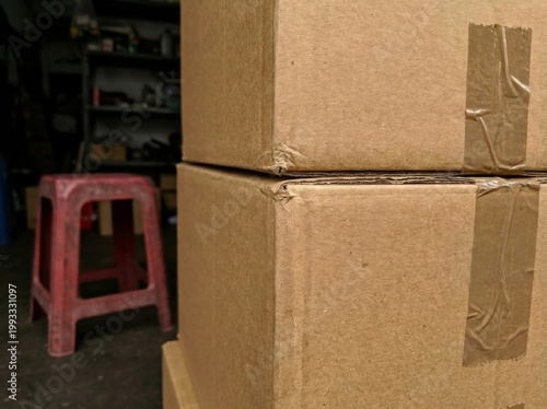 Two stacked cardboard boxes sit next to a red stool in a dimly lit storage area with shelves in the background.