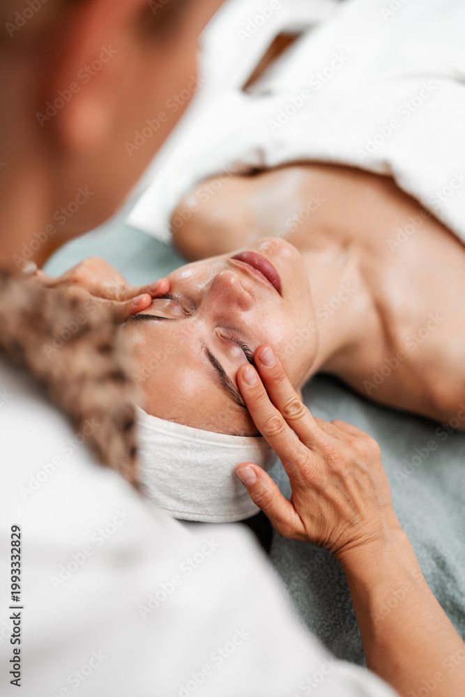 Fototapeta premium A professional aesthetician performs a relaxing facial massage on a young woman in a wellness spa. The client is lying down with eyes closed during the skincare treatment.