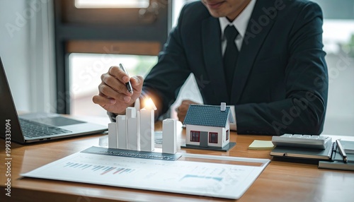 Man in suit analyzes property data with charts, pen, miniature house, and laptop
