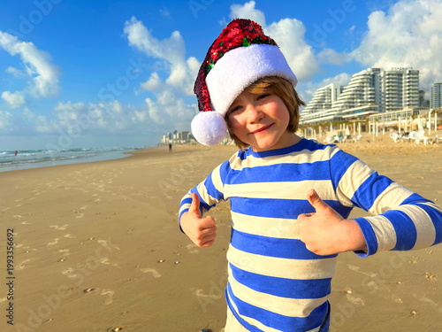 Christmas on the island! A boy in a Santa Claus hat on the beach by the sea. Winter holidays, family vacation