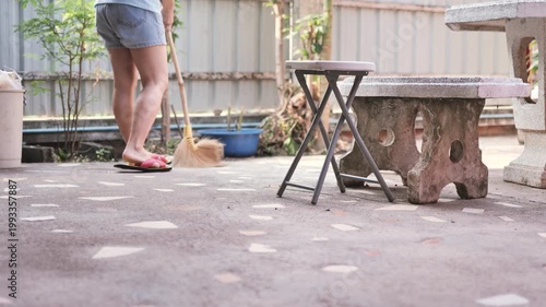 Cropped view of a person sweeping a tiled patio using a traditional straw broom in a home garden setting. The image represents daily chores, cleanliness, and simple lifestyle routines in a residential