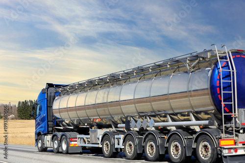 Blue truck pulling shiny tank semi-trailer at speed on road in golden light. Side view, copy space.