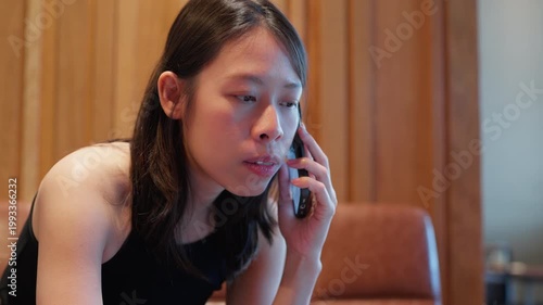 An Asian female professional communicates via smartphone in a modern wooden accented interior, representing the work from anywhere and digital nomad concept for business presentations.