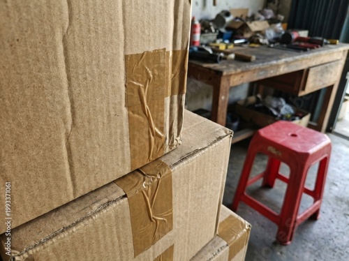 A cluttered workshop scene with stacked cardboard boxes in the foreground and a red stool next to a workbench laden with tools.