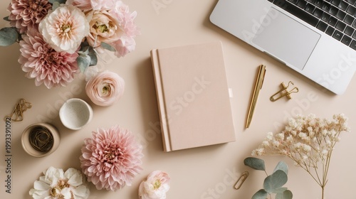 Overhead shot of a workspace. Laptop, book, pen, floral arrangement, stationery, and small cups rest on a flat, neutral surface