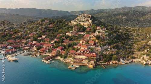 The historic fortress of Simena sits on a peak in the village of Kalekoy, Turkey. Aerial drone shot shows red-roofed houses by the sea in the village of Kalekoy, Turkey