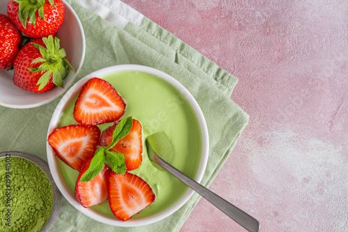 Matcha yogurt topped with fresh strawberries and mint leaves in bowl with a spoon. Top view with copy space. Healthy dessert, green smoothie breakfast on pastel pink background. Flat lay