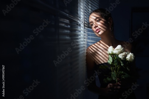 Woman Holds Red Roses While Standing Near a Window With Light Shining Through...