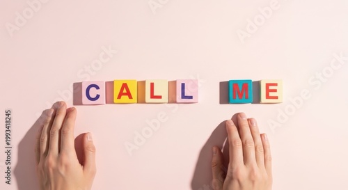 Hands frame colorful letter blocks spelling CALL ME on a pale pink surface, a playful, minimalist invitation that feels both flirty and sweetly nostalgic
