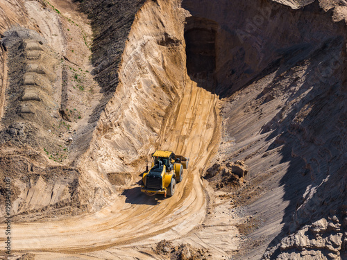 Aerial view shows a yellow wheel loader on a narrow winding haul road in an open pit mine, fresh tire tracks, layered sediment, and a shadowed adit under midday light.