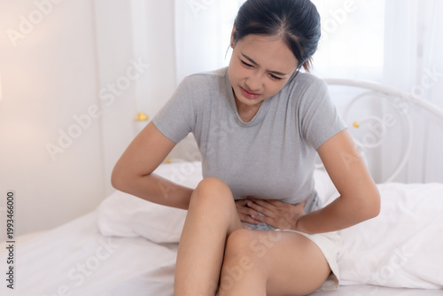 Tableau sur toile Asian women sitting on bed holding abdomen with pain, stomach ache or menstrual cramps, digestive problem concept, healthcare and illness, discomfort expression