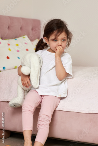 cute child girl sitting on her bed, holding a plush bunny, biting her fingers and looking away with a pensive expression